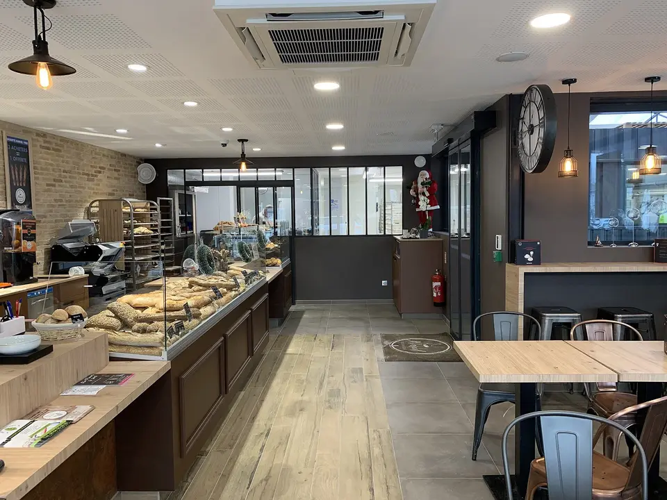 Intérieur d’une boulangerie : vitrines à pain, sol carrelé, tables et tabourets près de l’entrée